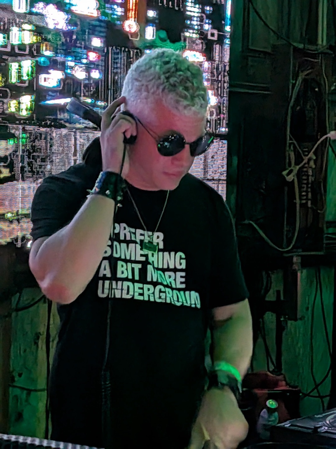 Dyson Hope DJing in a dark club, one hand holding headphones to his ear, wearing round dark sunglasses and a black T-shirt that reads "I prefer something a bit more underground," with a wall of glitching video screens behind him.